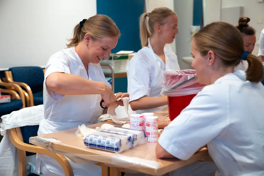 En kvinne klipper i en bandasje. Flere studenter i hvitt tøy sitter rundt henne.