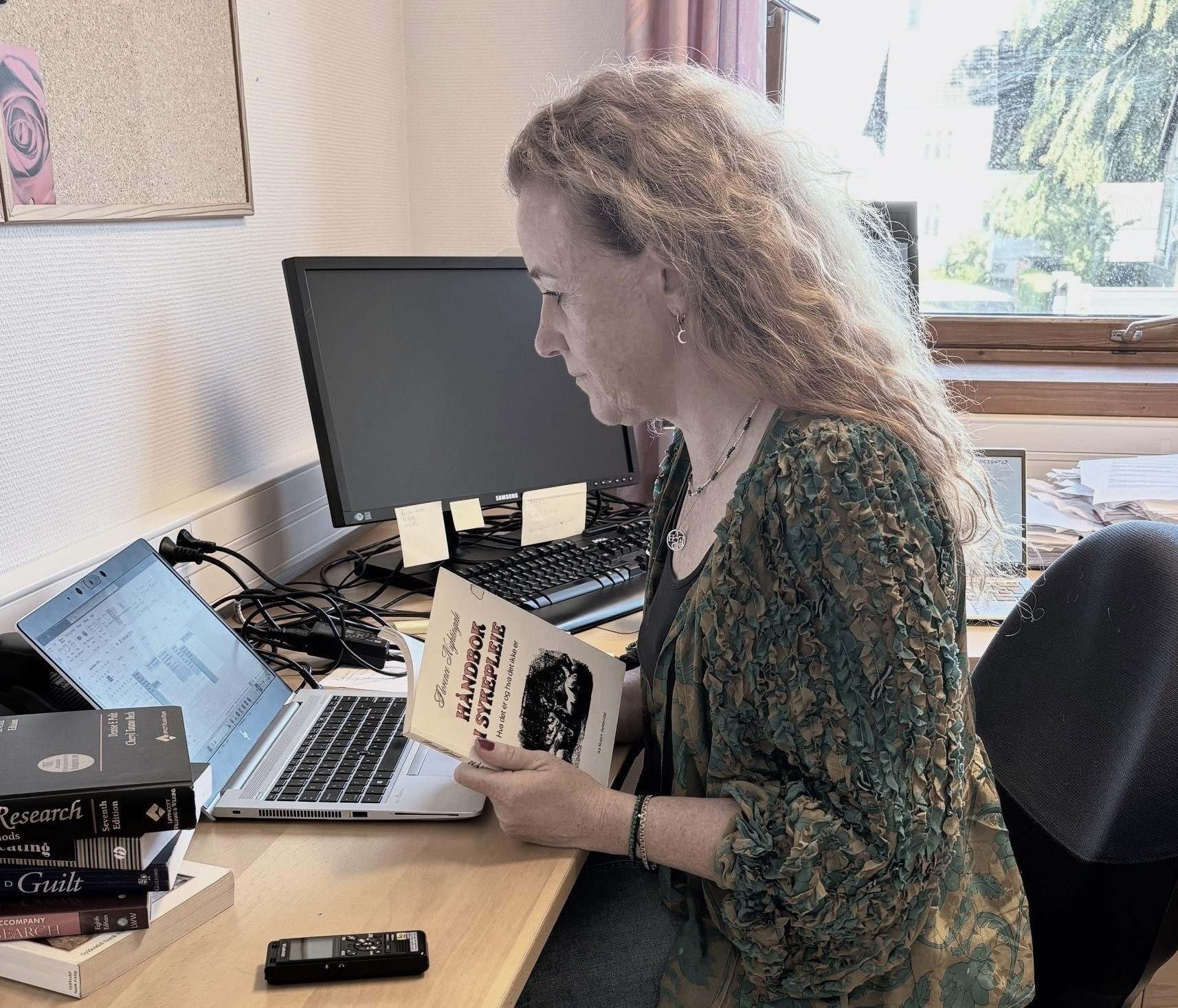 En forsker som ser på en bærbar datamaskin med håndbok i sykepleie i hendene