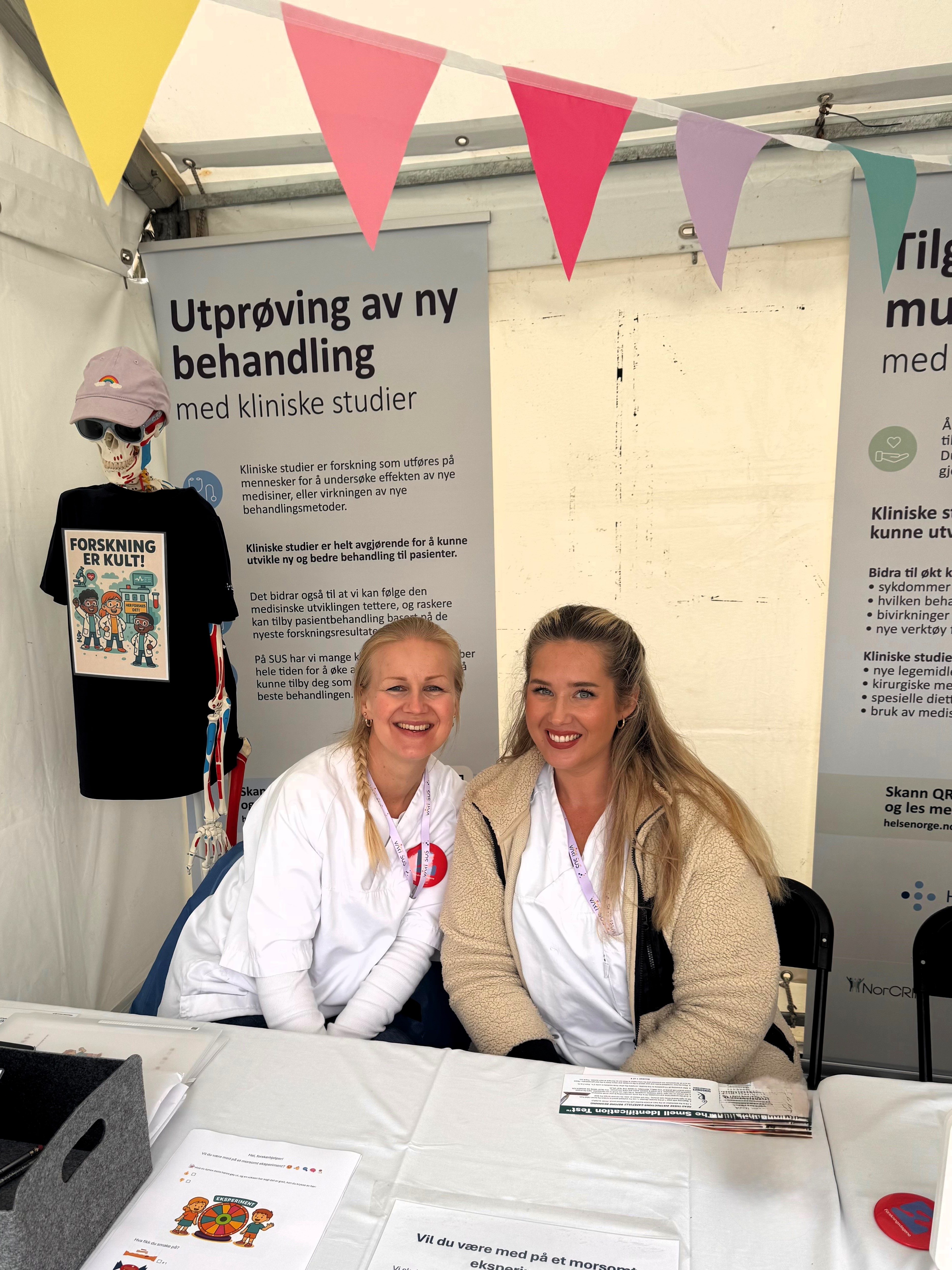 To sykepleiere som sitter ved et bord med et skilt bak seg