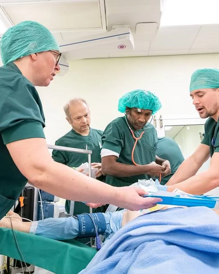 Simulering i akuttmottak. Tre helsepersonell står over en person som leker pasient. Foto.
