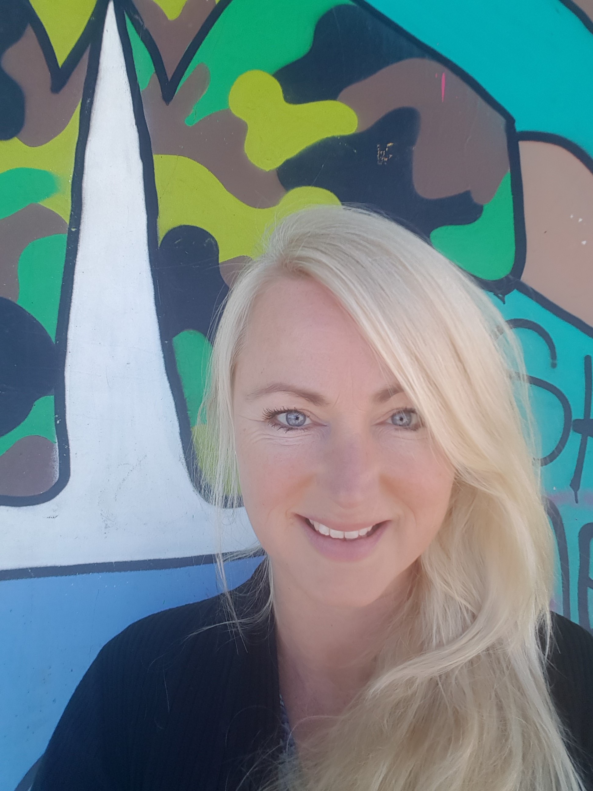 Kvinne med blondt hår står foran fargerik vegg.