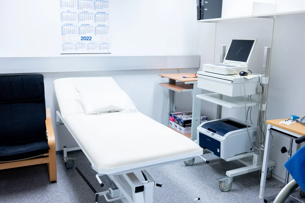 Foto av et undersøkelsesrom med stol, seng, apparat og benker.