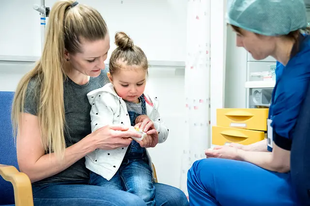 Barn på fanget til mor. Sykepleier med blå klær ser på dem. Foto.