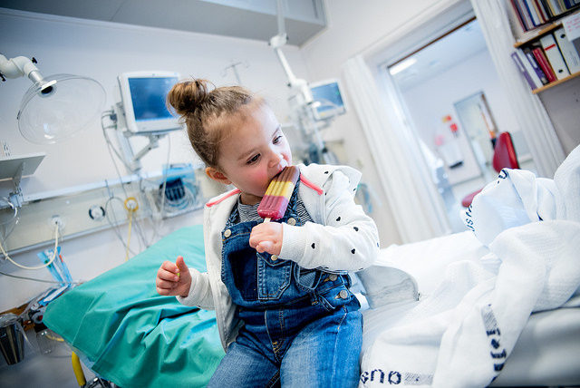 Barn som sitter på en sykehusseng og spiser is. Foto.