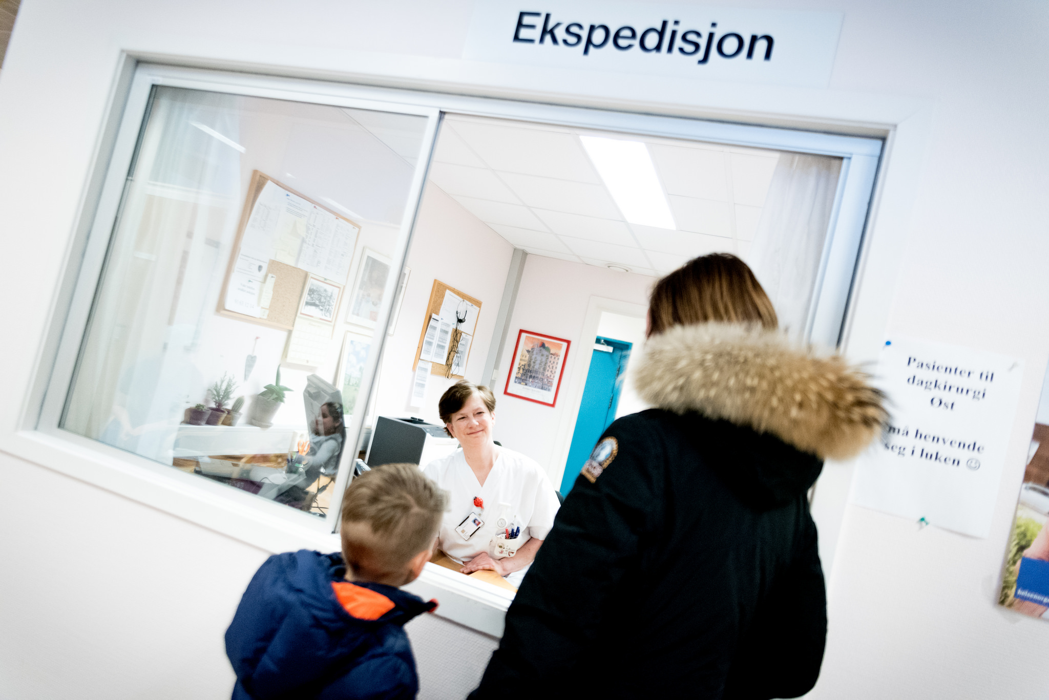 Mor og sønn snakker med sykepleier i hvite klær i ekspedisjonen. Foto.