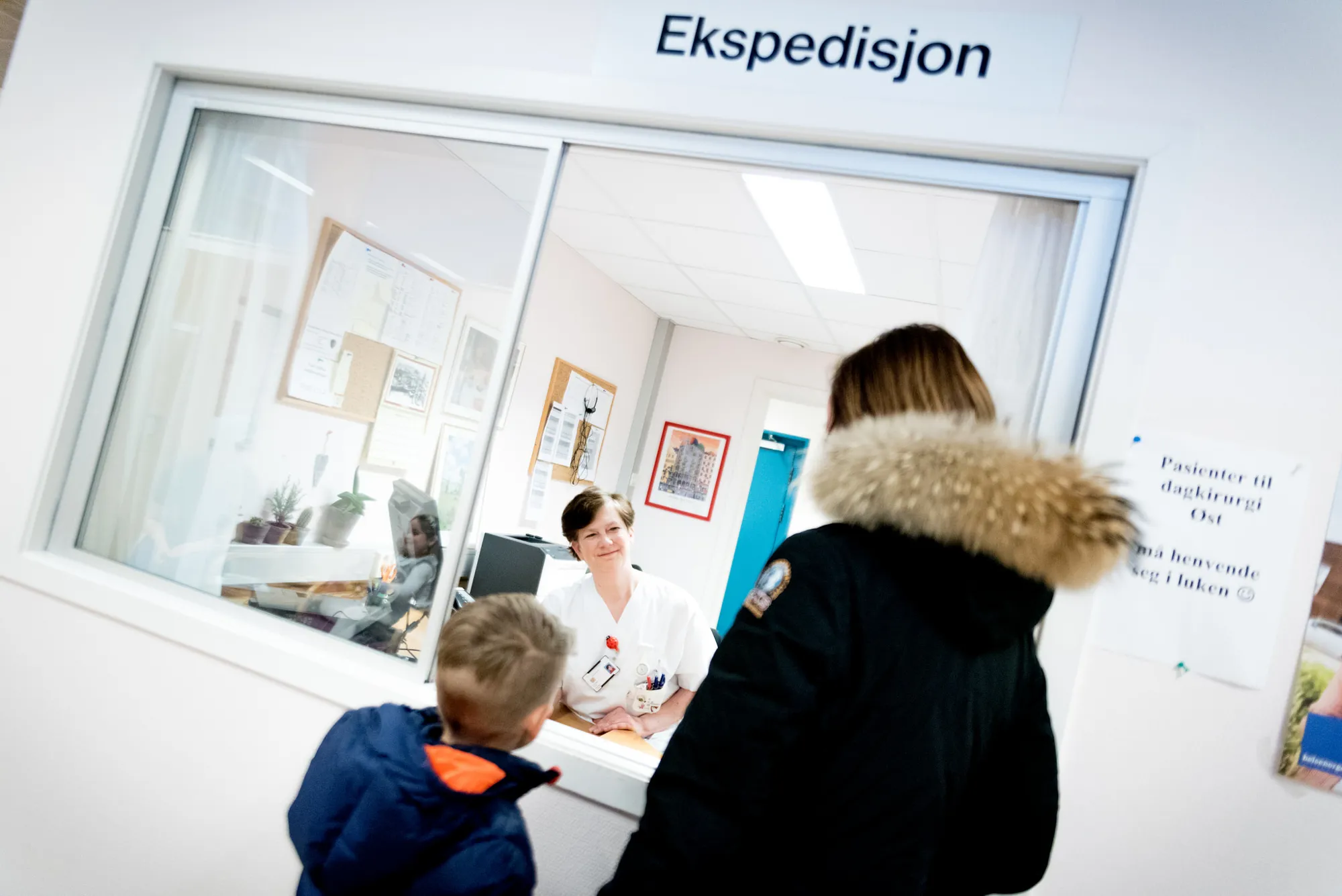 Mor og sønn snakker med sykepleier i hvite klær i ekspedisjonen. Foto.