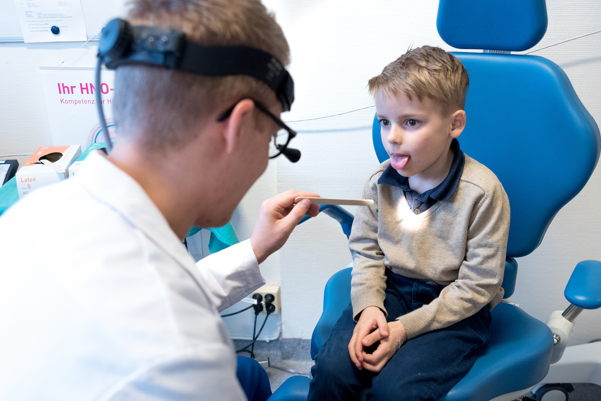 Liten gutt på en blå stol som strekker ut tungen. Lege med hodelykt ser inn i munnen. Foto.
