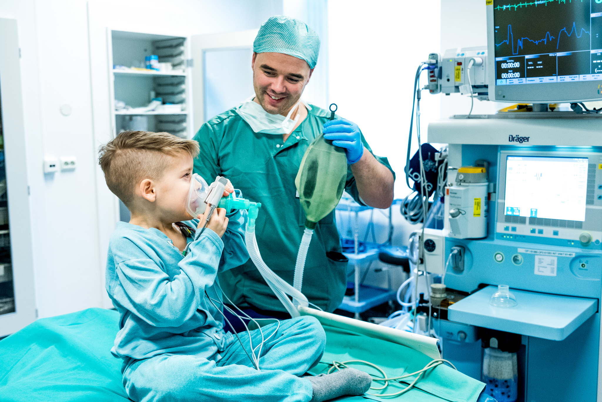 Liten gutt i sykehusklær sitter på en operasjonsseng og tester en pustemaske. Anestesilege hjelper til. Foto.