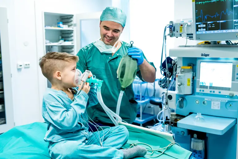 Liten gutt i sykehusklær sitter på en operasjonsseng og tester en pustemaske. Anestesilege hjelper til. Foto.