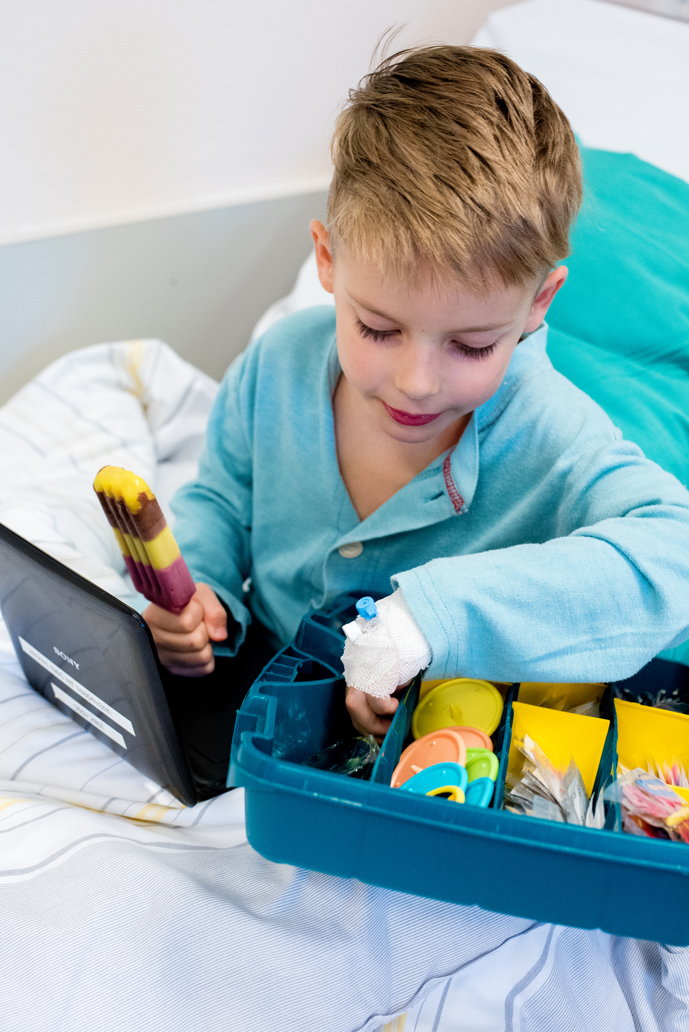 Liten gutt i sykehusklær sitter i en sykehusseng og spiser is. Han ser på små ting i en boks.