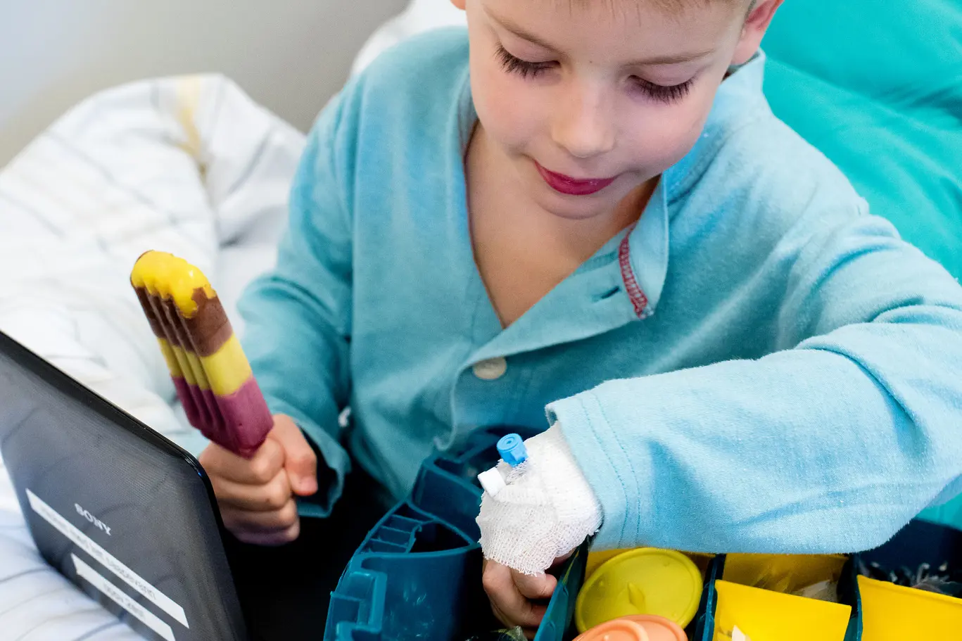 Liten gutt i sykehusklær sitter i en sykehusseng og spiser is. Han ser på små ting i en boks.