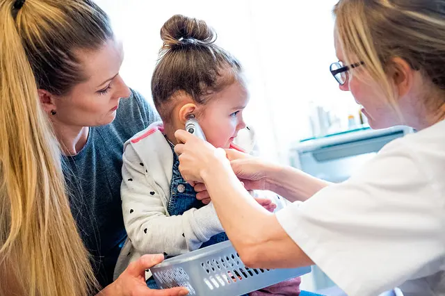 Barn som sitter på fanget til mor og har temperaturmåler i øret. Sykepleier holder måleren.