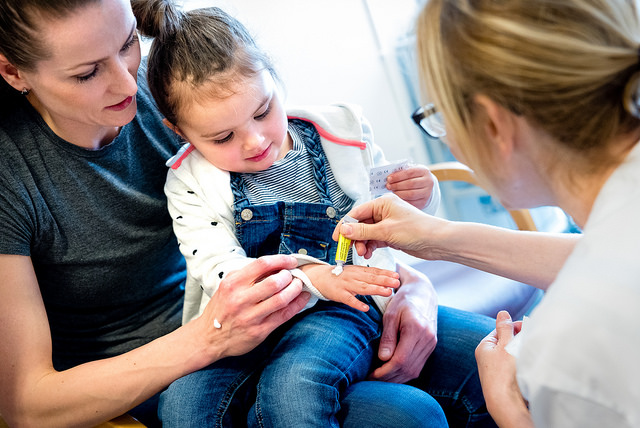 Barn som sitter på fanget til mor og får krem på hånden av en sykepleier. Foto.