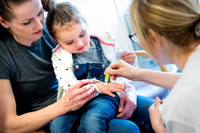 Barn som sitter på fanget til mor og får krem på hånden av en sykepleier. Foto.