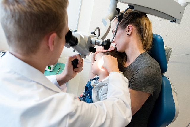 Lege ser inn å ørene på et barn. Barnet sitter på fanget til mor. Foto.