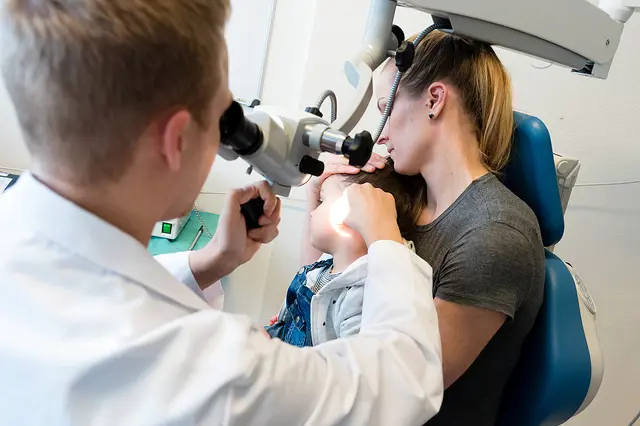 Lege ser inn å ørene på et barn. Barnet sitter på fanget til mor. Foto.