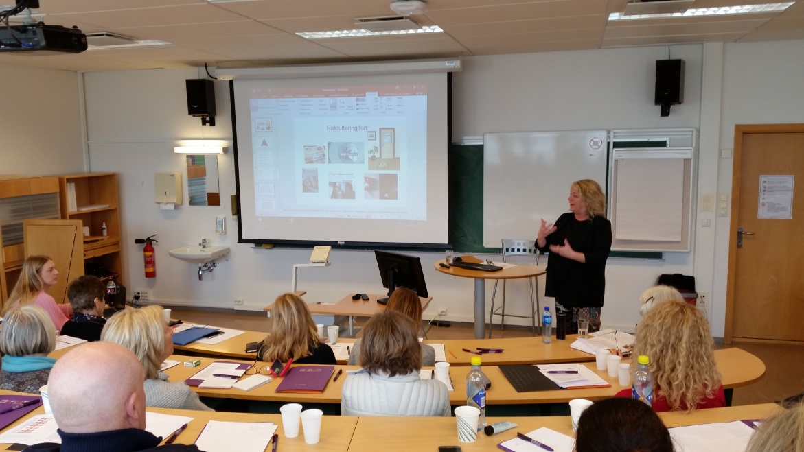 En kvinne står og snakker foran en liten forsamling av mennesker.