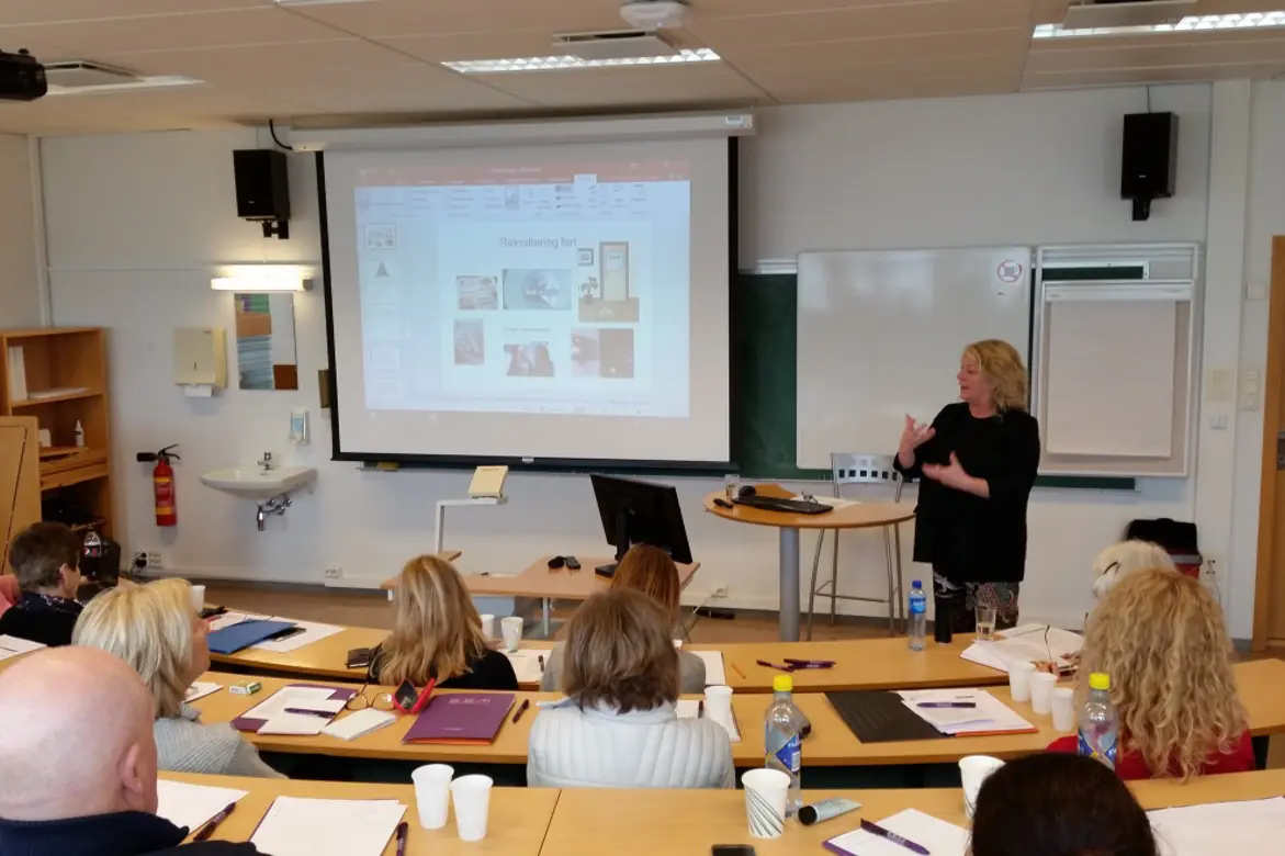 En kvinne står og snakker foran en liten forsamling av mennesker.