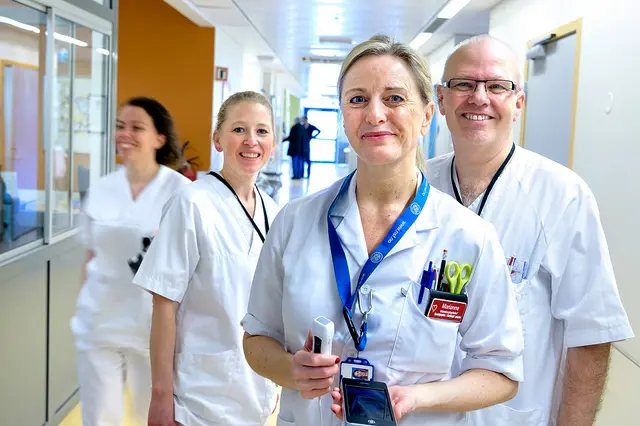 Fire hvitkledde personer - tre kvinner og en mann - ser inn i kamera og smiler. Hun ene holder et lite ultralydapparat i hendene