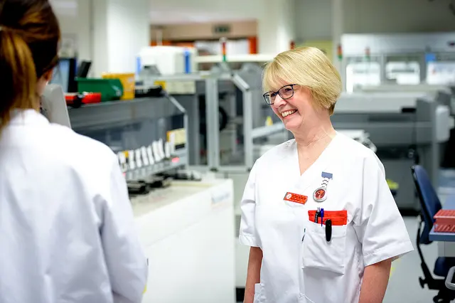 To kolleger ser på hverandre - den ene har ryggen til, den andre er vendt mot kamera og smiler.